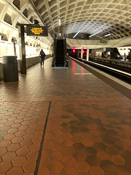 A picture of an upwards escalator on a train platform. The escalator is in the background of the picture in the center. On the left side there is a half wall along with a trash can and a information display, while on the right side of the image there are the train tracks.